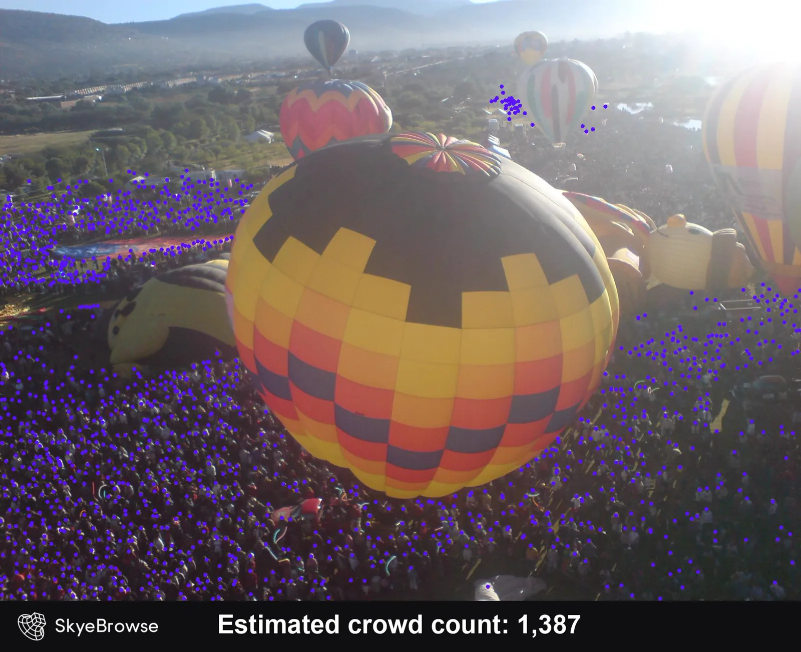 Aerial view of hot-air balloons inflating above a packed field of spectators marked with purple dots