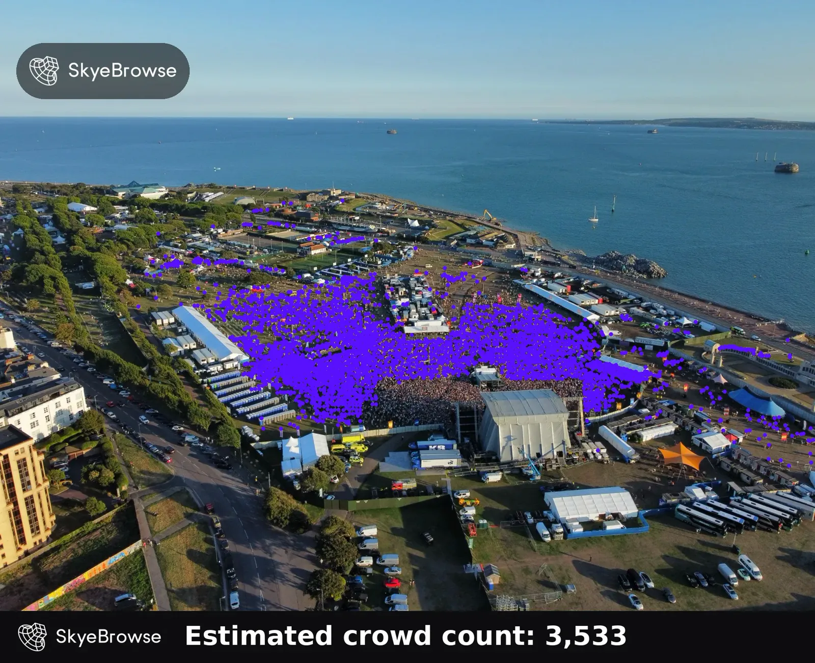 Drone view of a seaside music festival with the crowd in front of the stage covered in purple head markers